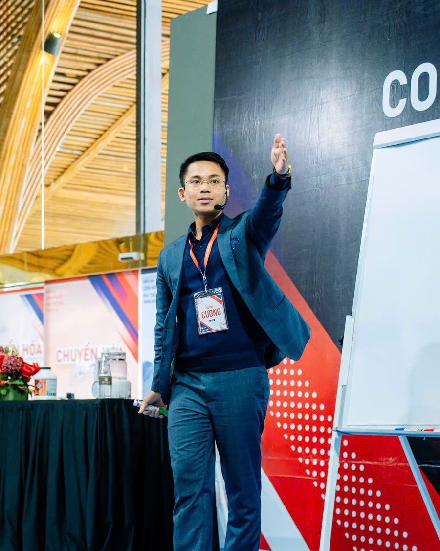 Lê Văn Cương đứng lớp, đeo micro cài tai và thẻ tên, một tay chỉ lên cao