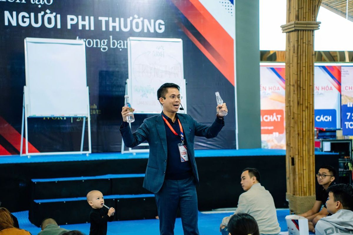 Lê Văn Cương cầm hai chai nước minh hoạ trước học viên, phía sau là backdrop Người phi thường trong bạn