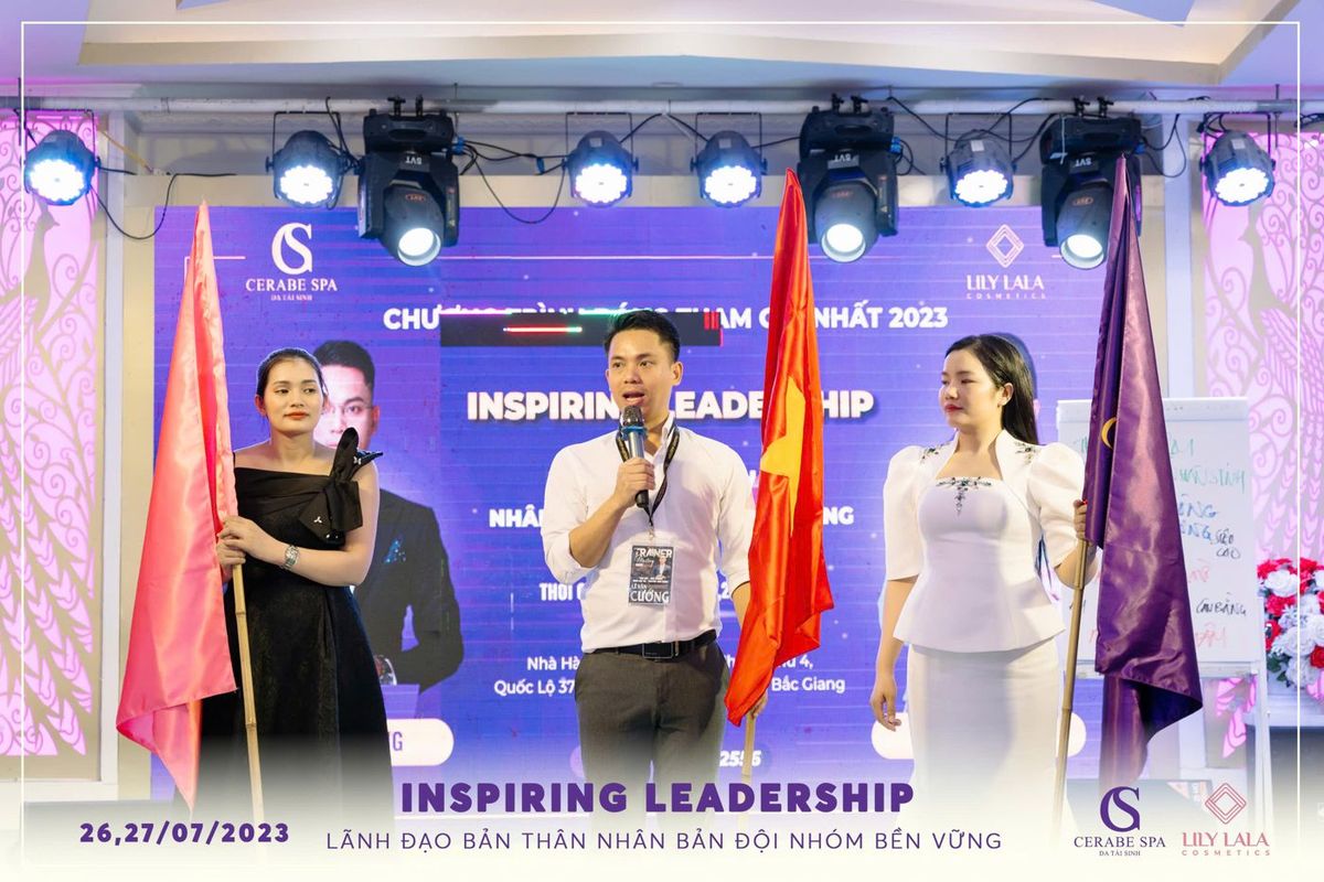 Lê Văn Cương cầm micro trên sân khấu Inspiring Leadership ngày 26–27/07/2023, hai bên là cờ