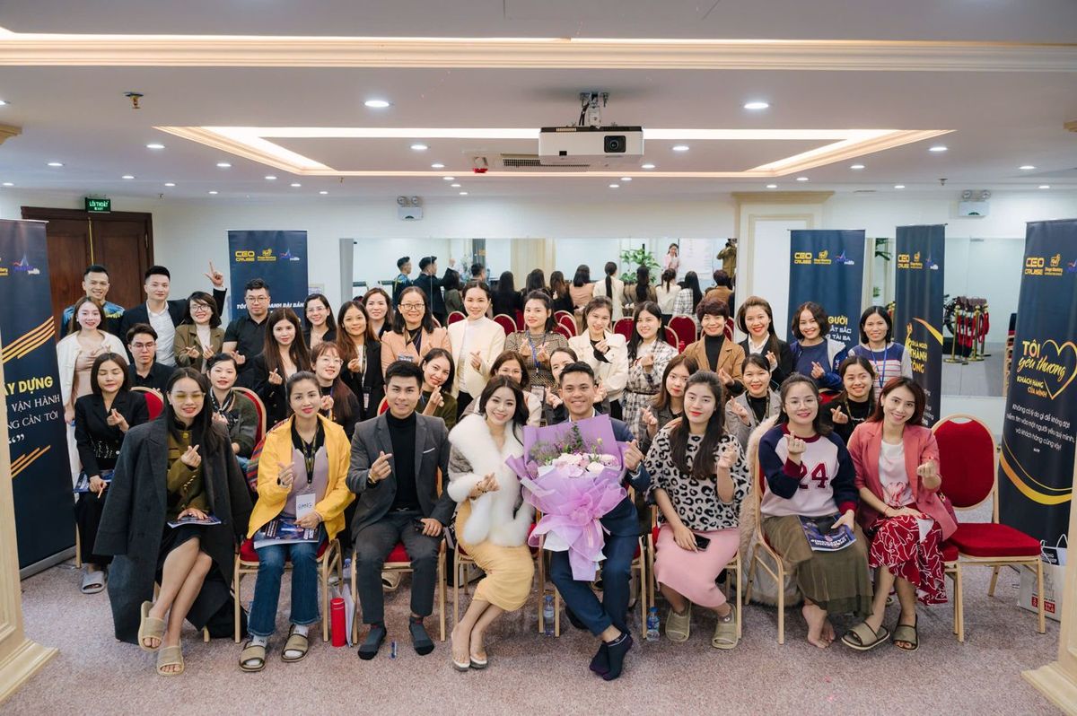 Lê Văn Cương ôm bó hoa, ngồi chụp ảnh cùng các học viên trong phòng học có standee Stage Mastery
