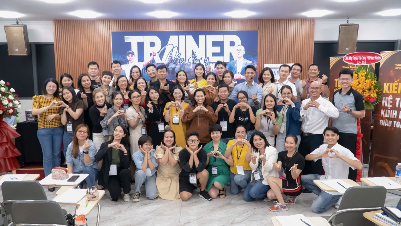 Học viên lớp Trainer Mastery chụp ảnh tập thể, nhiều người làm động tác trái tim trước backdrop Trainer Mastery