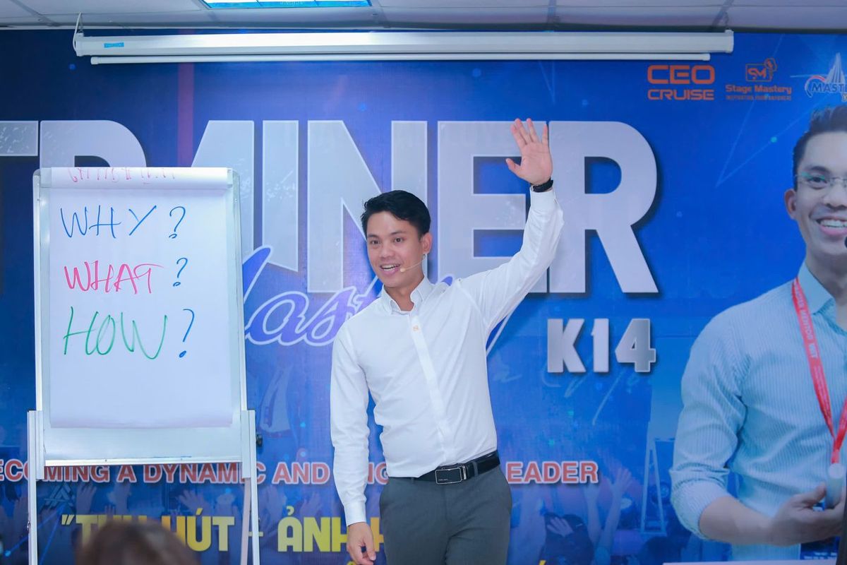 Lê Văn Cương giơ tay đứng lớp trước backdrop Trainer Mastery K14, bên cạnh bảng lật ghi Why, What, How